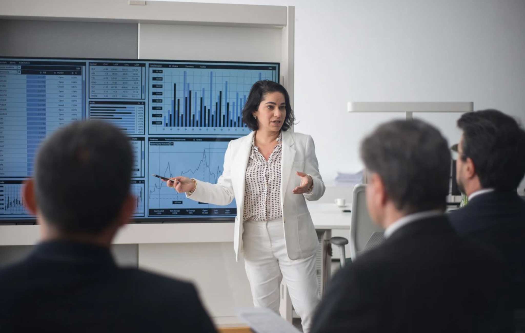 A woman in a business suit presents confidently to an engaged audience in a professional setting.